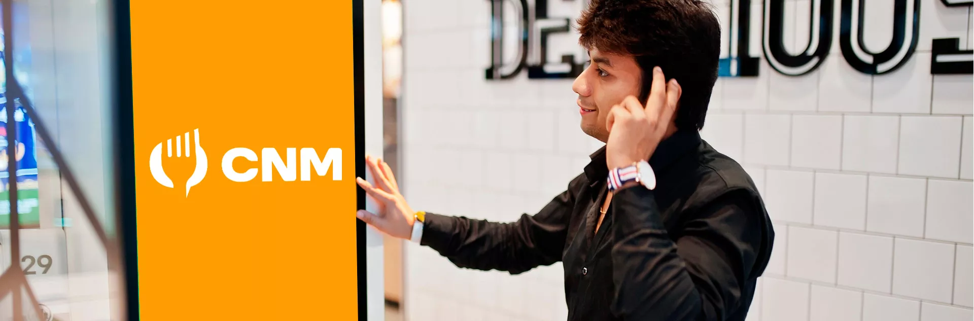 Homem de camisa preta e relógio branco sorrindo enquanto interage com um totem de autoatendimento laranja com o logotipo do CNM, em frente a uma parede branca de azulejos.